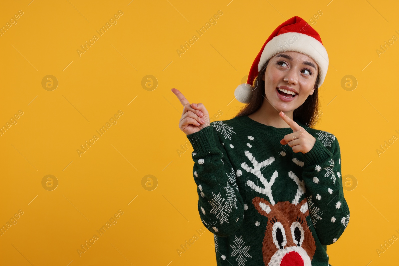 Photo of Happy young woman in Christmas sweater and Santa hat pointing at something on orange background. Space for text