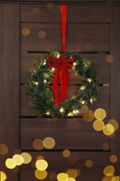Beautiful Christmas wreath with red bow and festive lights hanging on door indoors