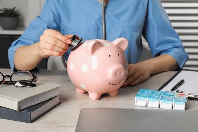 Doctor with stethoscope and piggy bank at wooden table in hospital, closeup. Medical insurance