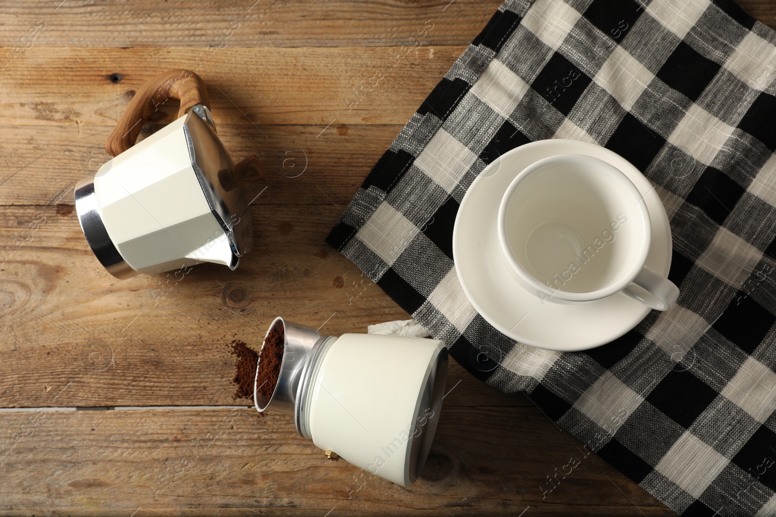 Photo of Moka pot, cup with saucer, ground coffee and checkered towel on wooden table, flat lay