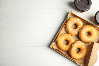 Delicious donuts in box and coffee on white table, flat lay. Space for text