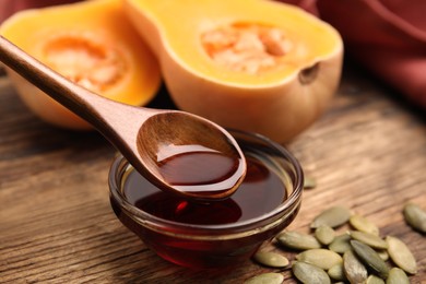 Photo of Spoon with pumpkin oil over bowl and seeds on wooden table, closeup