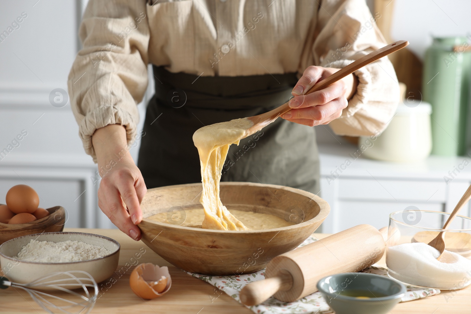 Photo of Woman kneading dough with spoon in bowl at wooden table in kitchen, closeup