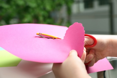 Photo of Child cutting out paper heart with craft scissors at table indoors, closeup