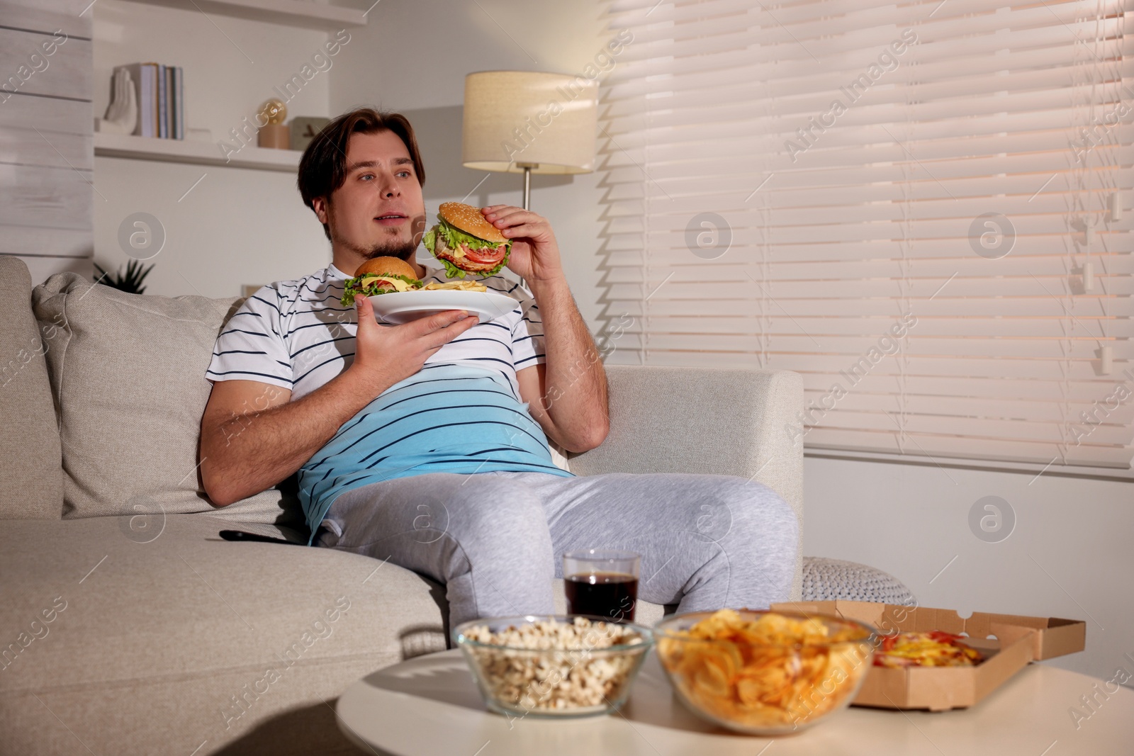 Photo of Overweight man with plate of burgers and French fries on sofa at home