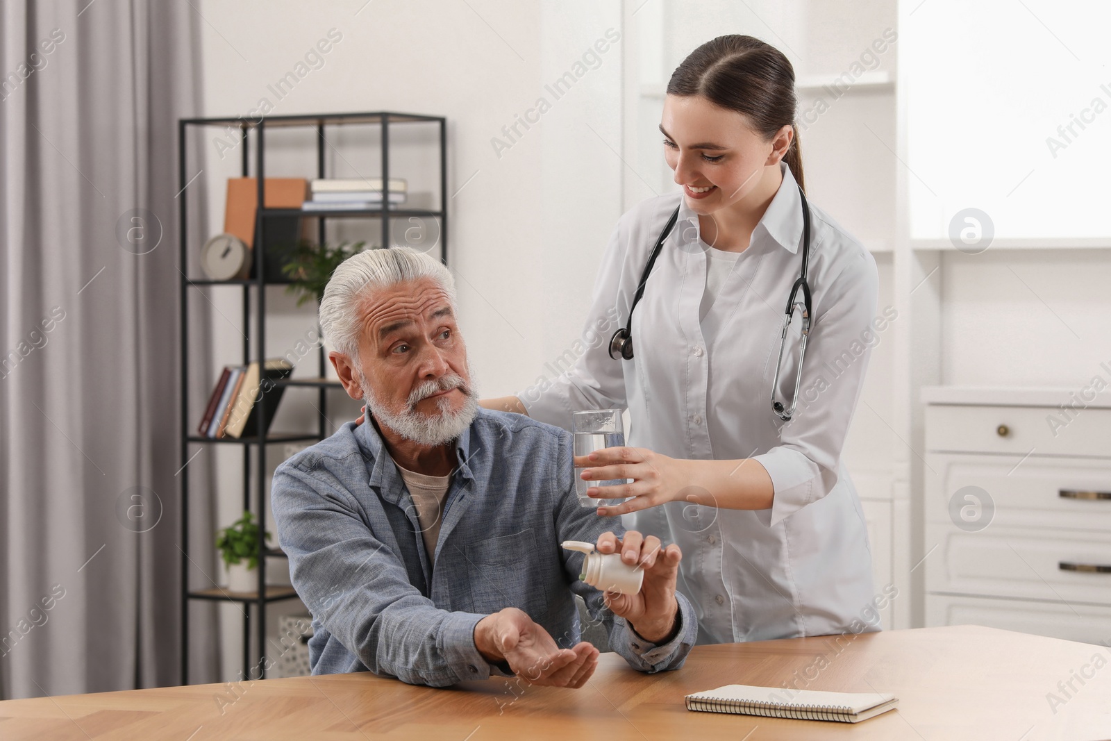 Photo of Young healthcare worker giving glass of water to senior man with pills at wooden table indoors