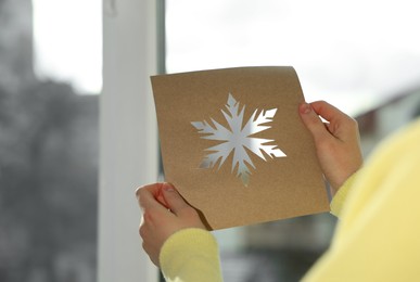 Photo of Woman holding snowflake stencil near window at home, closeup