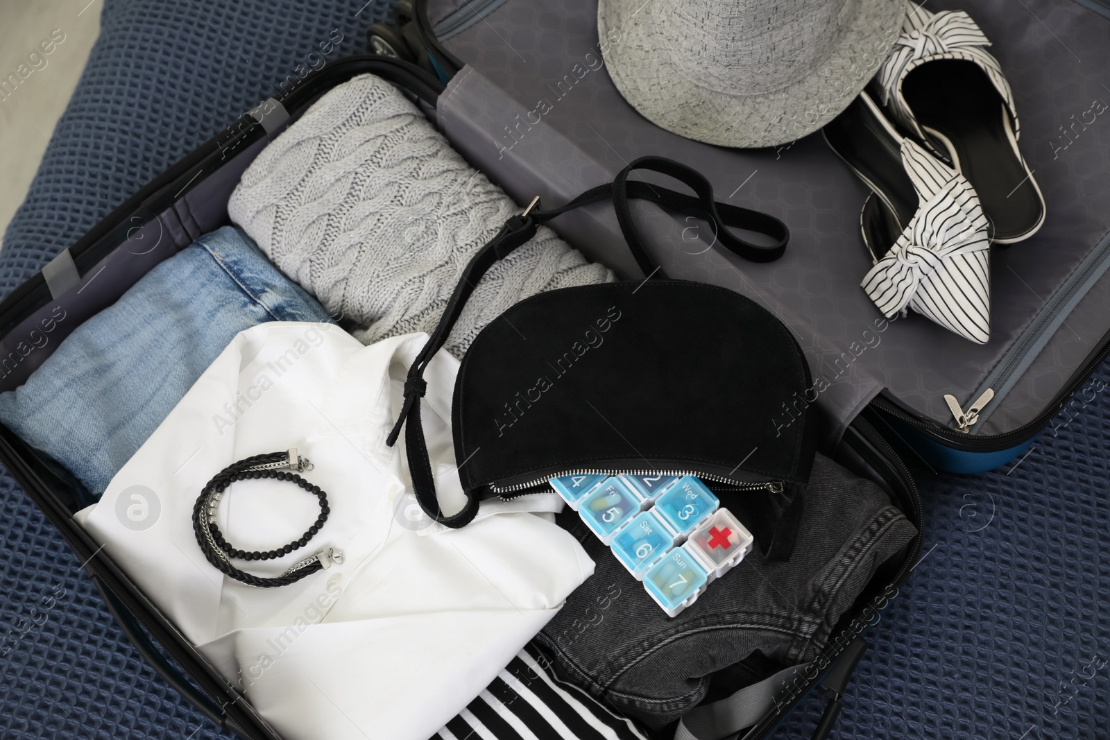 Photo of Open suitcase with packed clothes, accessories and pill box on blue blanket, above view