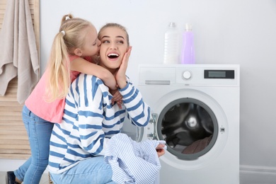 Little girl hugging her mother while she doing laundry at home