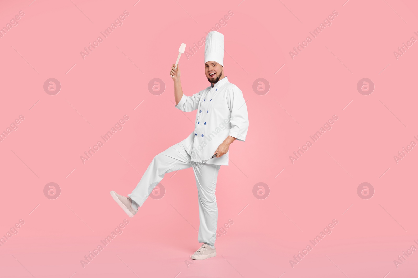 Photo of Happy professional confectioner in uniform holding spatula and whisk on pink background