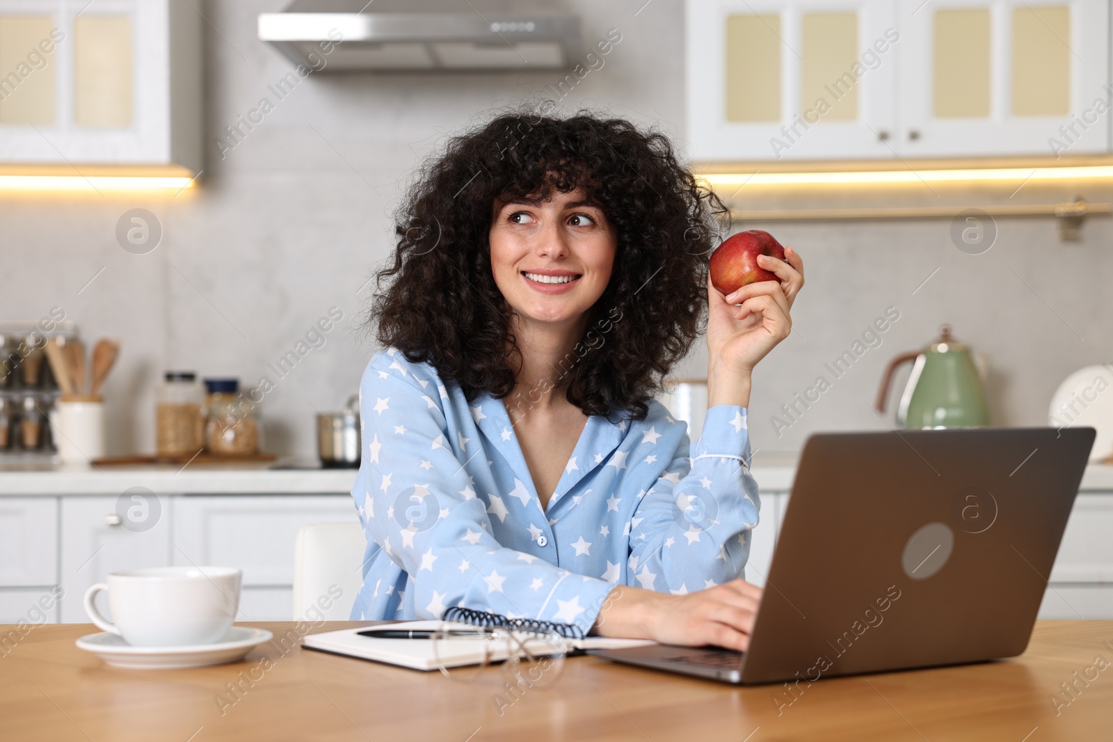 Photo of Beautiful young woman in stylish pyjama with apple using laptop at wooden table in kitchen