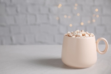 Cup of delicious hot cocoa with marshmallows on white table against blurred lights, space for text