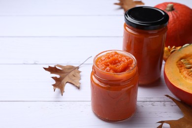 Jars of delicious pumpkin jam, fresh pumpkin and dry leaves on white wooden table, space for text