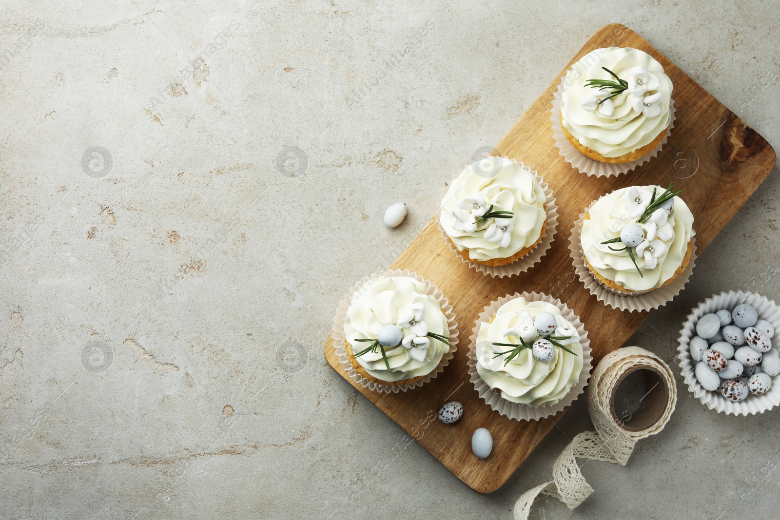 Photo of Tasty Easter cupcakes with vanilla cream, candies and ribbon on gray table, flat lay. Space for text