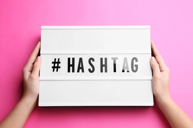 Photo of Woman holding light box with symbol and word Hashtag on pink background, top view