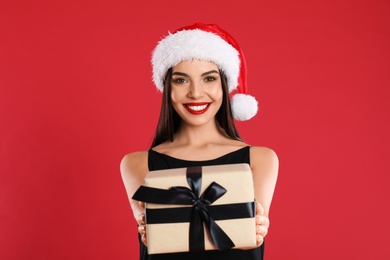 Photo of Woman in black dress and Santa hat holding Christmas gift on red background