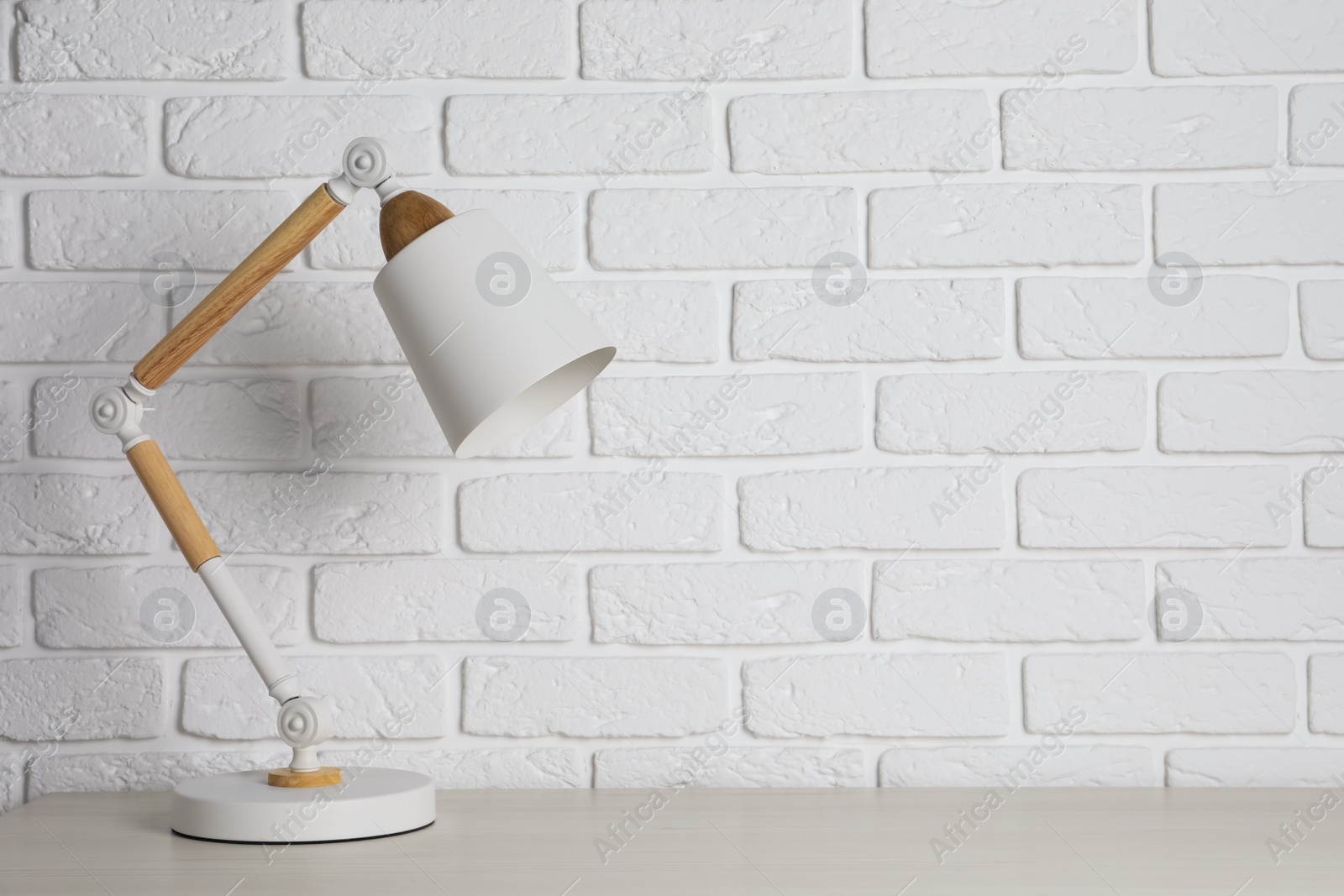 Photo of Stylish modern desk lamp on white wooden table near brick wall, space for text