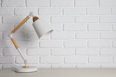 Photo of Stylish modern desk lamp on white wooden table near brick wall, space for text