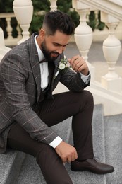 Young groom in elegant suit with beautiful boutonniere sitting on stairs outdoors