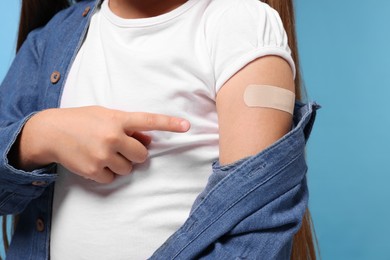Photo of Girl pointing at sticking plaster after vaccination on her arm against light blue background, closeup