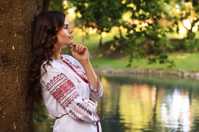 Photo of Beautiful woman in embroidered shirt near lake, space for text. Ukrainian national clothes