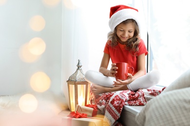 Photo of Cute little child in Santa hat with cup of cocoa sitting on windowsill at home. Christmas celebration