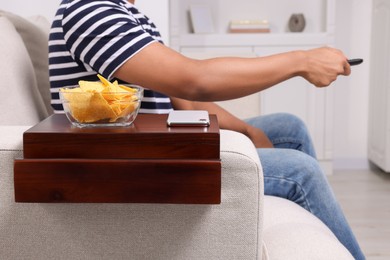 Photo of Nacho chips and smartphone on sofa armrest wooden table. Man holding remote control at home, closeup