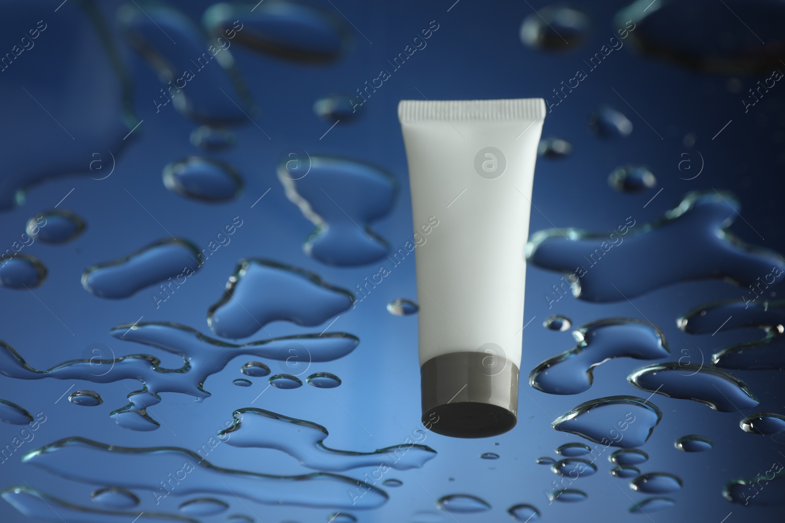 Photo of Moisturizing cream in tube on glass with water drops against blue background, low angle view. Space for text