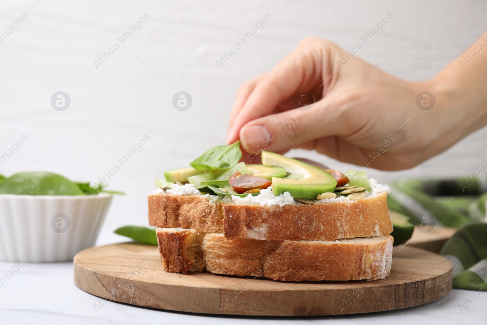 Photo of Woman putting spinach leaf on tasty vegan sandwich with avocado and tomato at white marble table, closeup