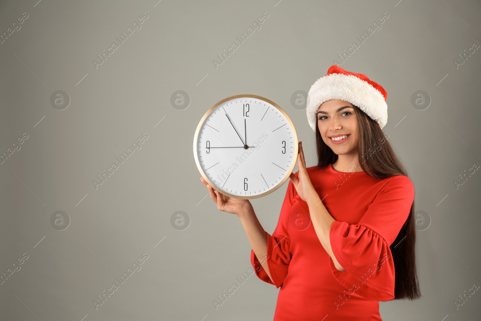 Photo of Young beautiful woman in Santa hat holding big clock on grey background. Christmas celebration