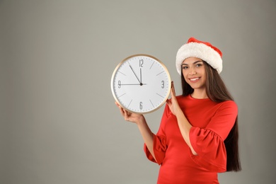 Young beautiful woman in Santa hat holding big clock on grey background. Christmas celebration