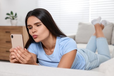 Photo of Young woman having video chat via smartphone and sending air kiss on sofa in living room