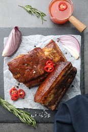 Photo of Pieces of baked pork belly served with sauce, rosemary and chili pepper on grey table, top view