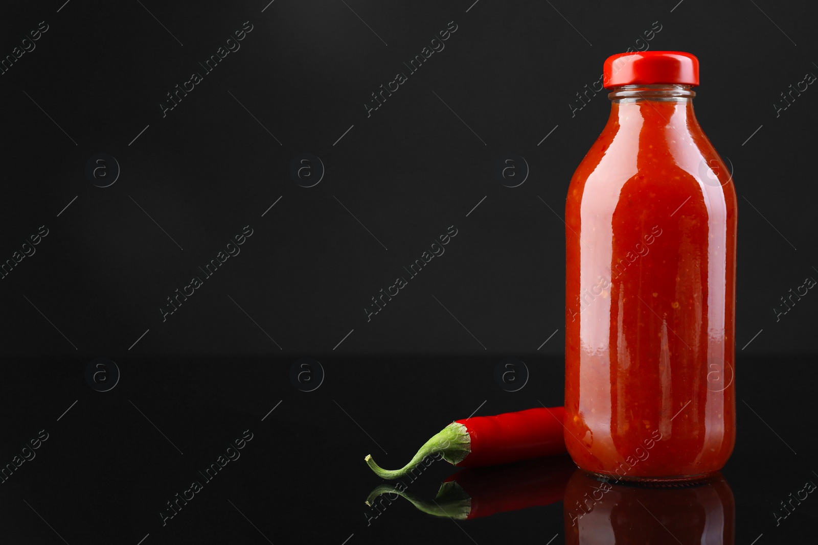 Photo of Spicy chili sauce in bottle and pepper against dark background, space for text