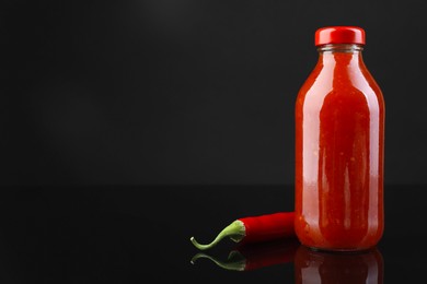 Photo of Spicy chili sauce in bottle and pepper against dark background, space for text