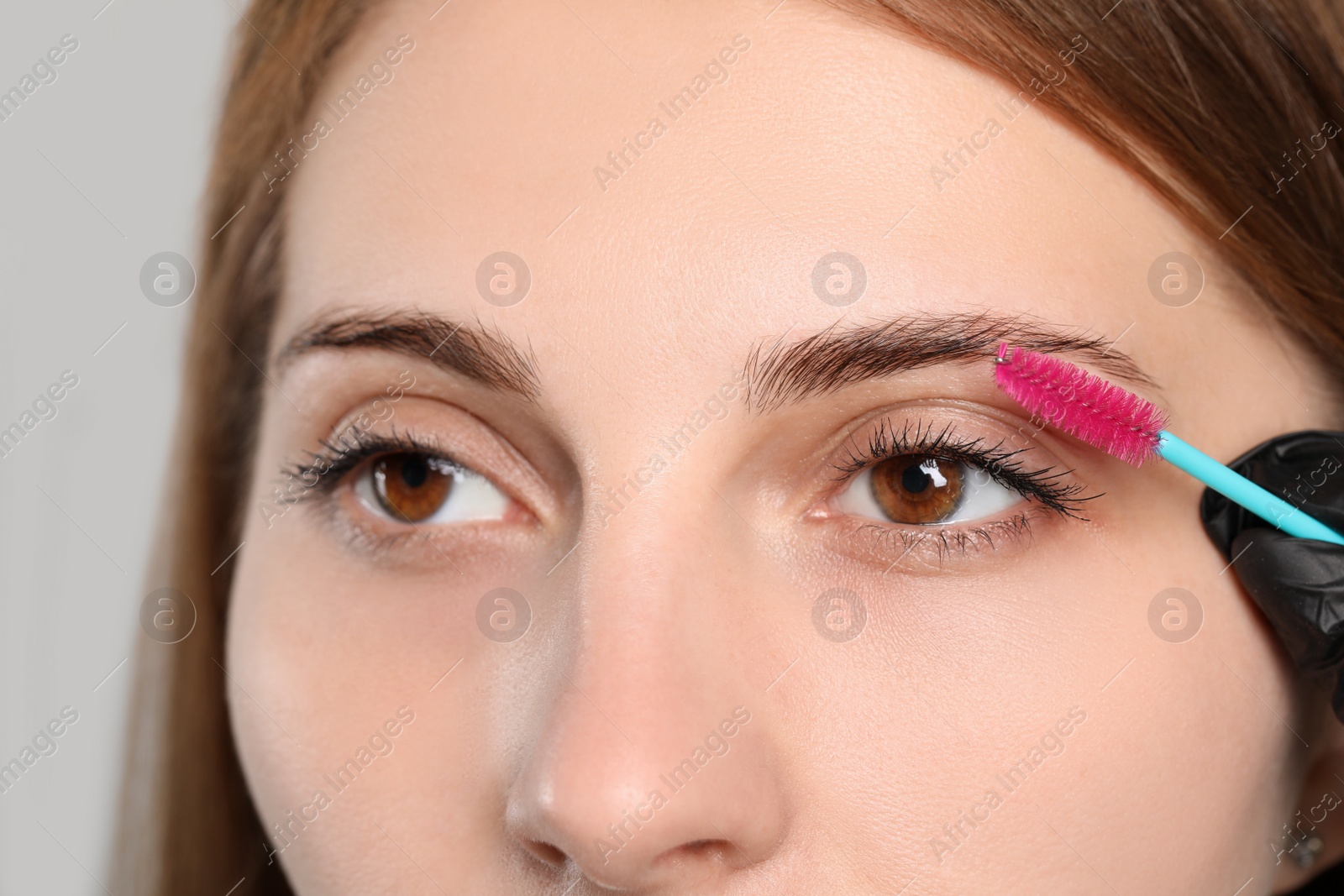 Photo of Beautician brushing woman's eyebrows before tinting on grey background, closeup