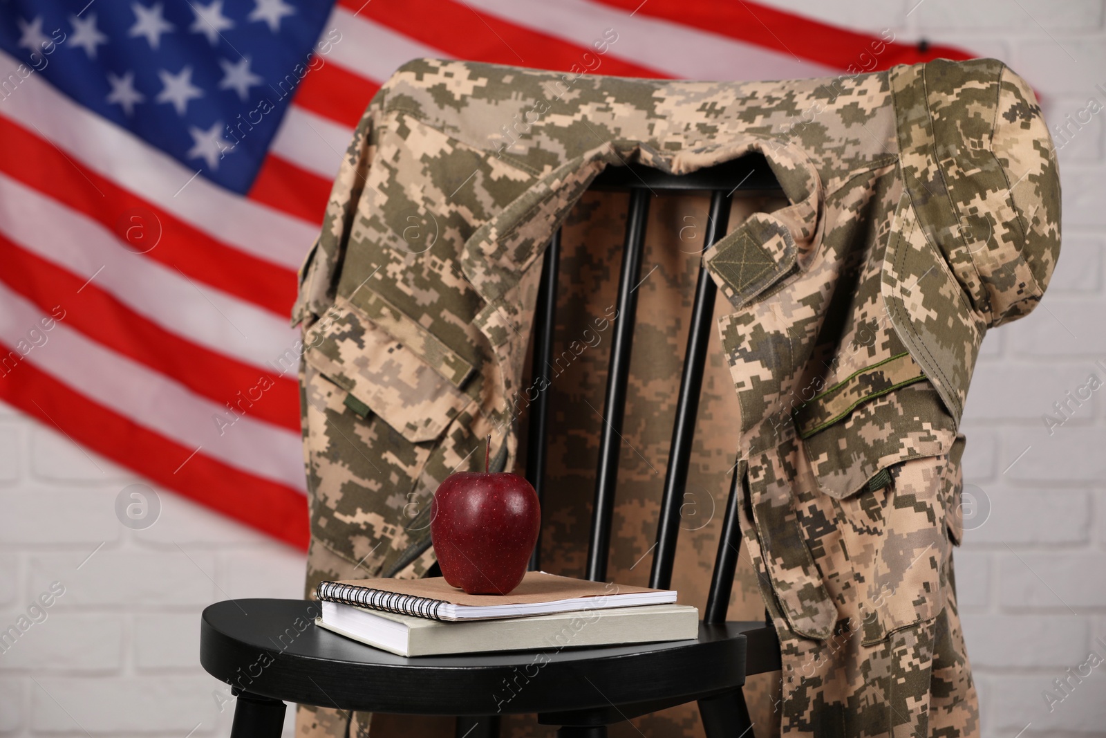 Photo of Soldier uniform, notebooks and apple on chair near flag of United States. Military education