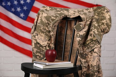 Soldier uniform, notebooks and apple on chair near flag of United States. Military education