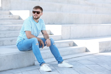 Photo of Young hipster man in stylish jeans sitting on stairs outdoors