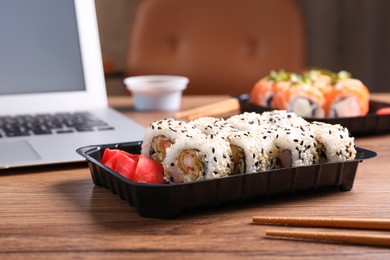 Photo of Tasty sushi rolls with shrimps in box near laptop on wooden table indoors
