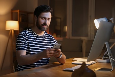 Home workplace. Man using smartphone near computer at wooden desk in room at night