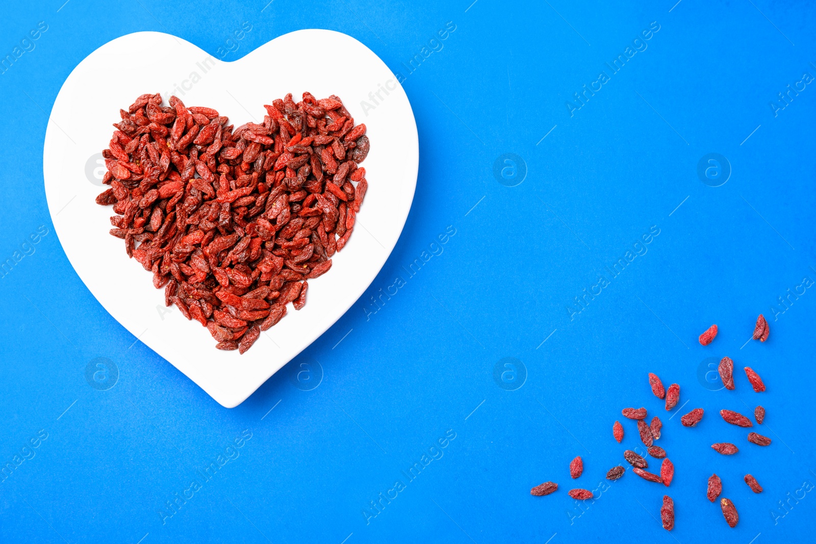 Photo of Dried goji berries in heart shaped plate on blue background, flat lay