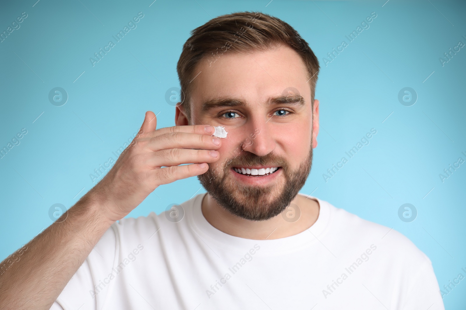 Photo of Happy young man applying facial cream on light blue background