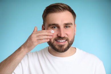 Photo of Happy young man applying facial cream on light blue background