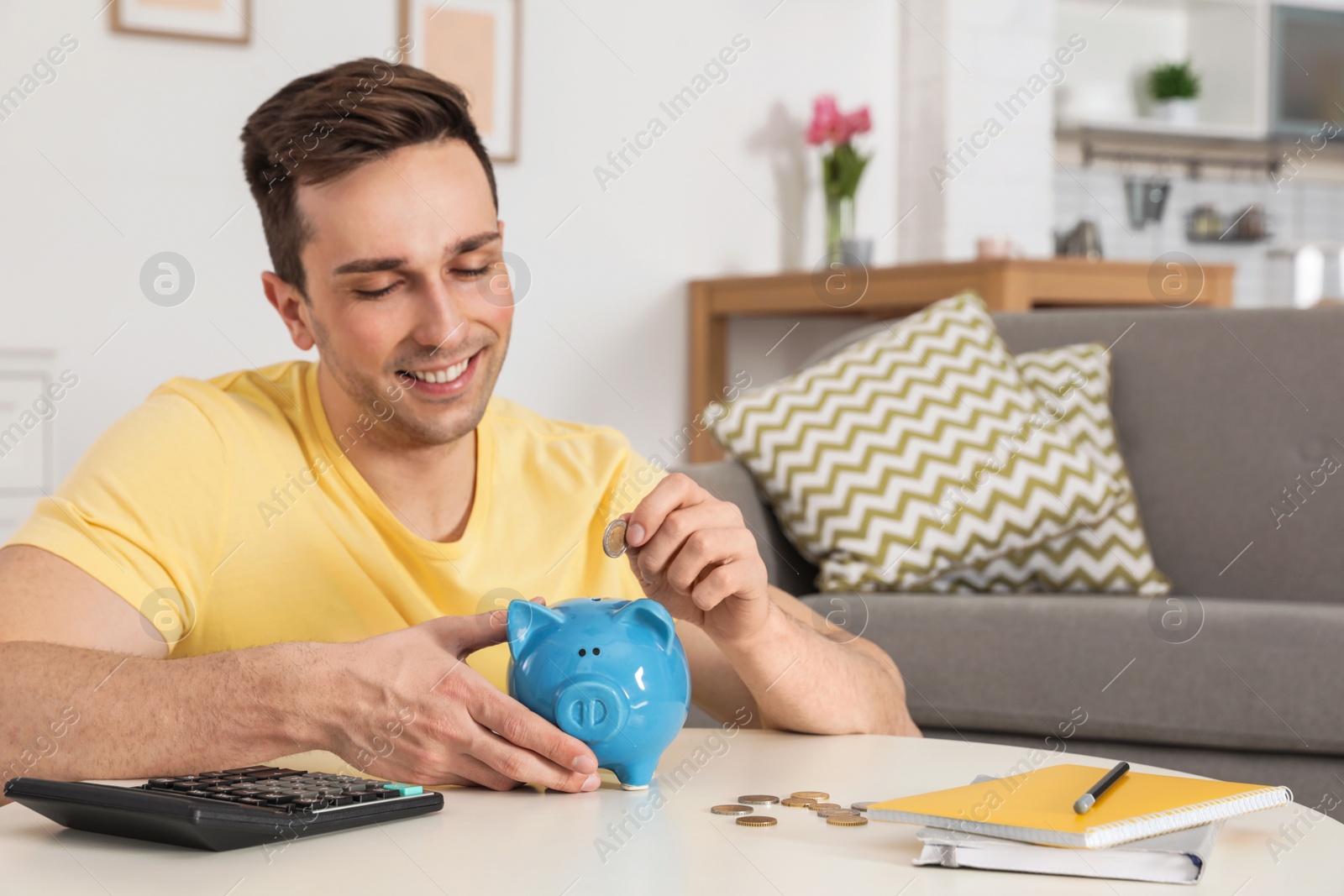 Photo of Happy man putting coin into piggy bank at table in living room. Saving money