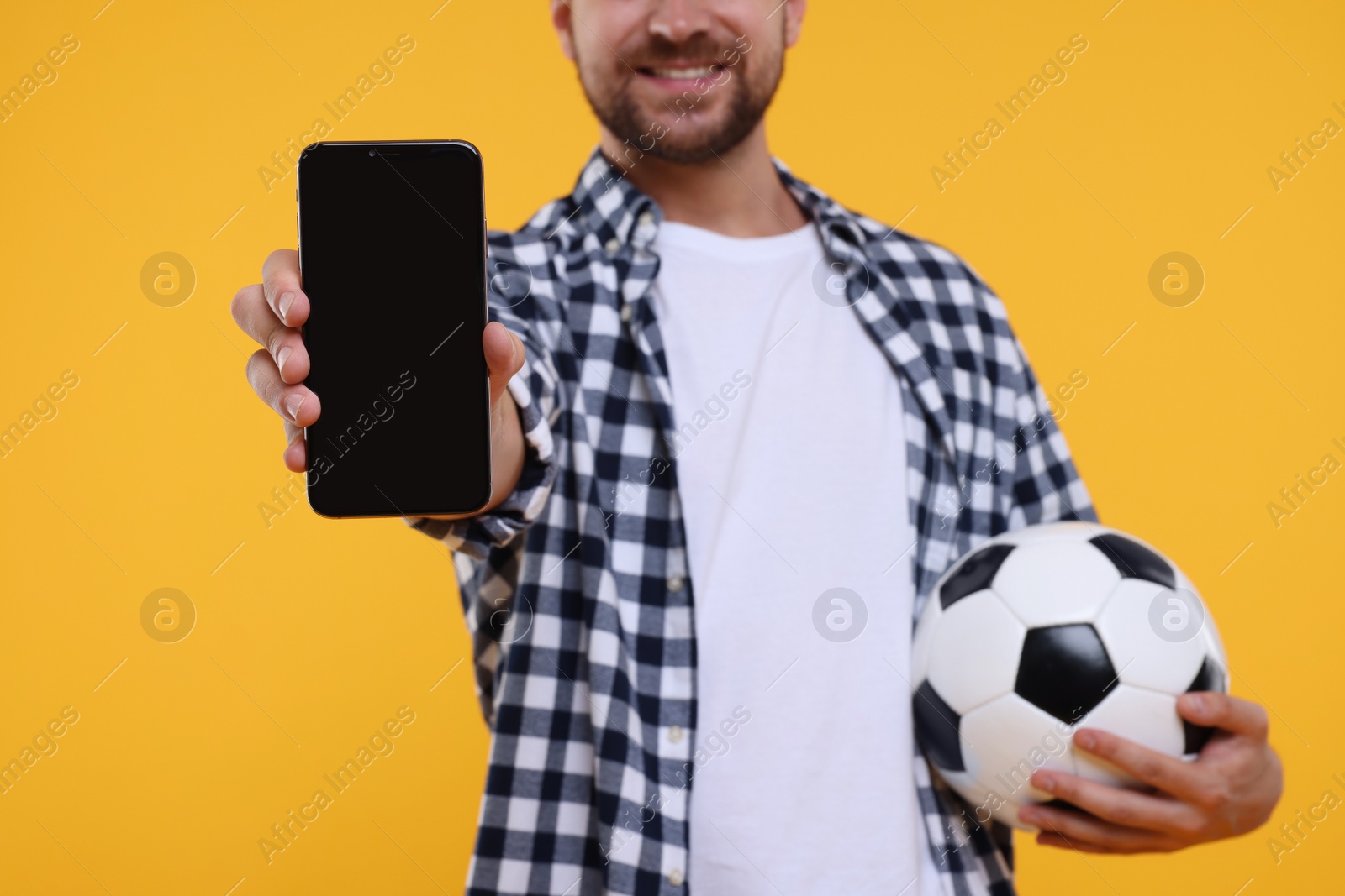 Photo of Sports fan with ball and smartphone on yellow background, closeup