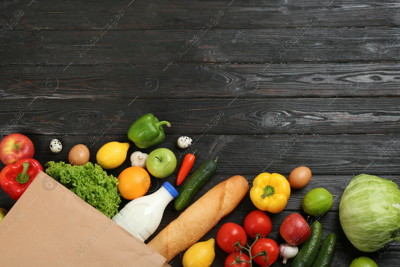 Photo of Flat lay composition with overturned paper bag and groceries on black wooden background. Space for text