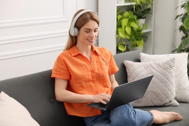 Woman in headphones using laptop on sofa near beautiful potted houseplants at home