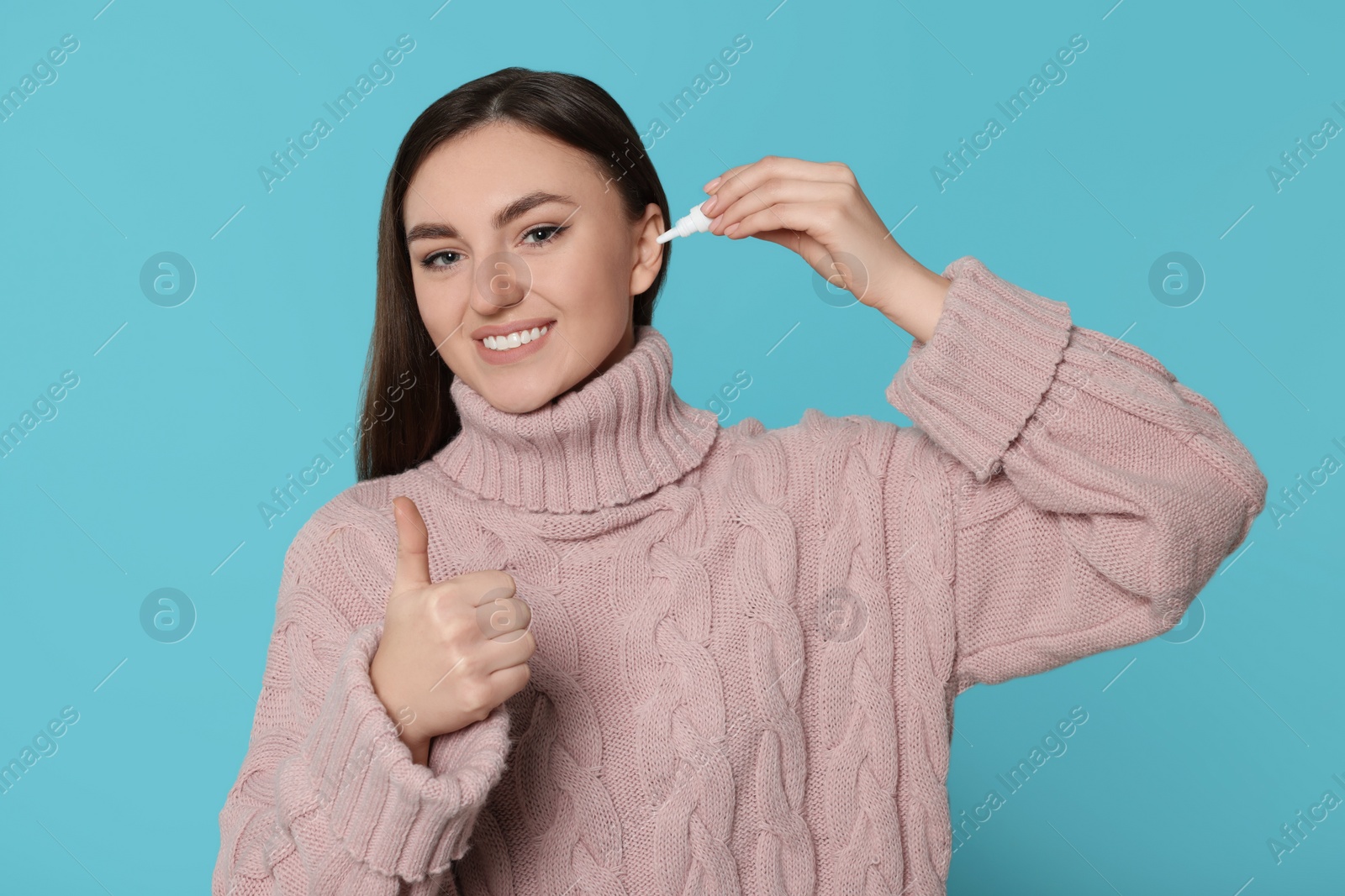 Photo of Young woman using ear drops and showing thumbs up on light blue background