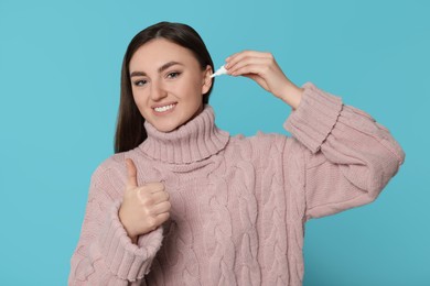 Young woman using ear drops and showing thumbs up on light blue background